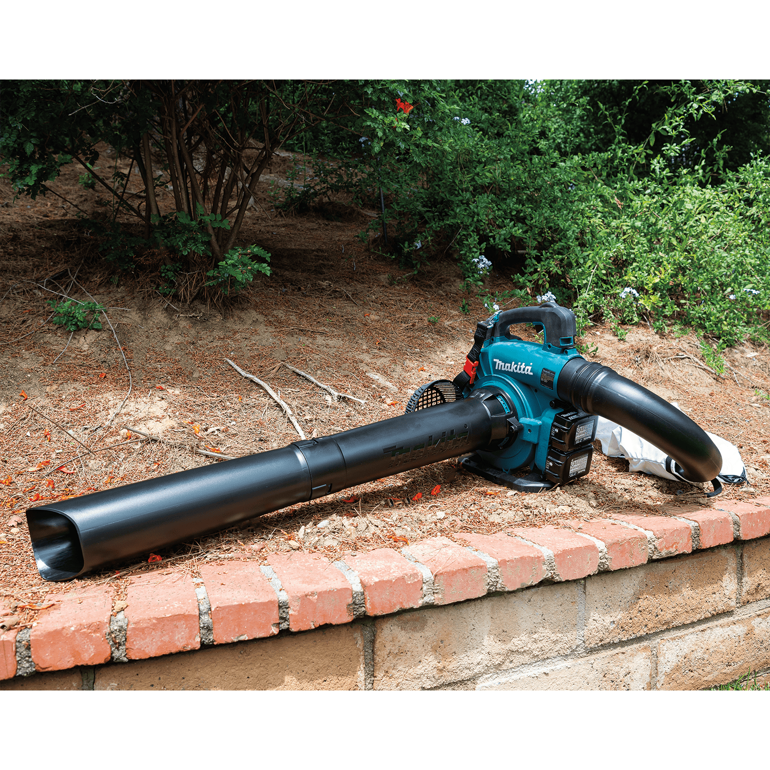 Makita XBU04PTV set up in vacuum and mulcher configuration resting near garden landscaping and brick wall.