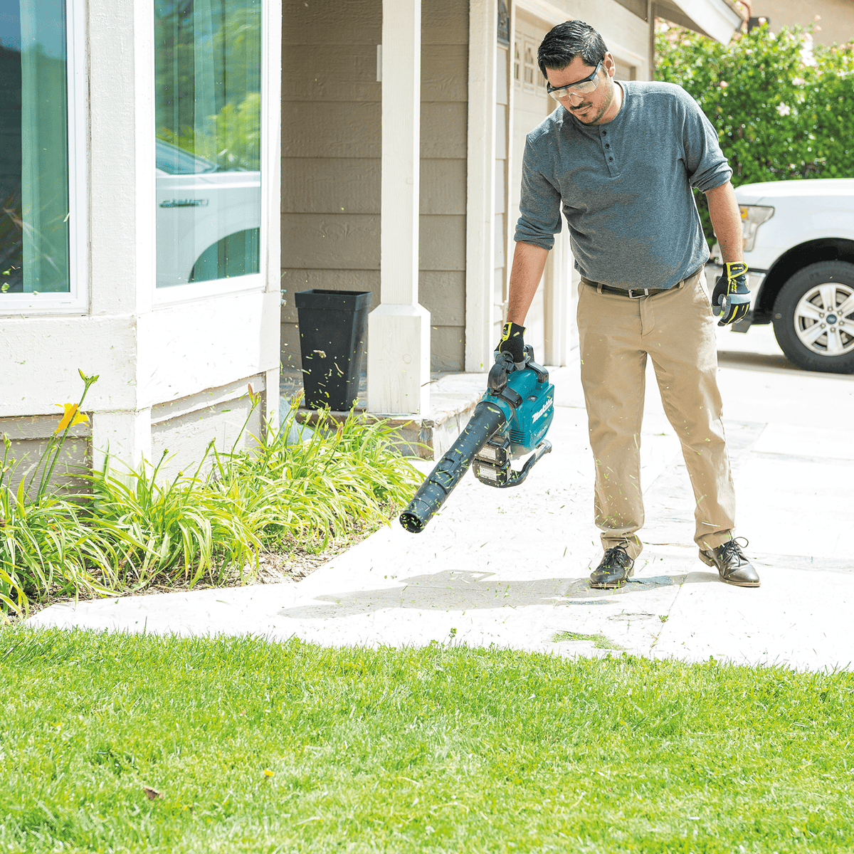 Worker clearing grass clippings and debris off a residential concrete driveway with a Makita blower.