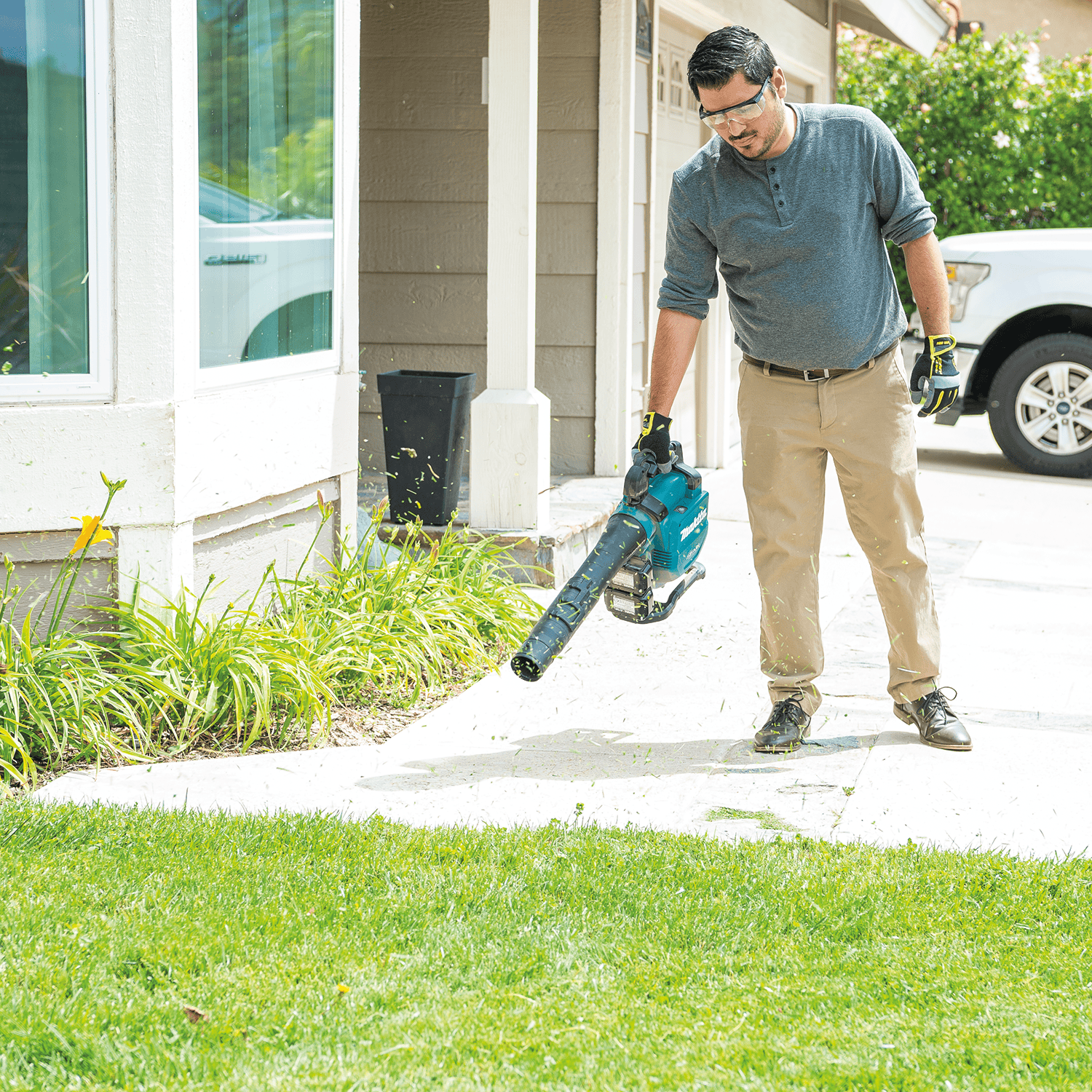 Worker clearing grass clippings and debris off a residential concrete driveway with a Makita blower.