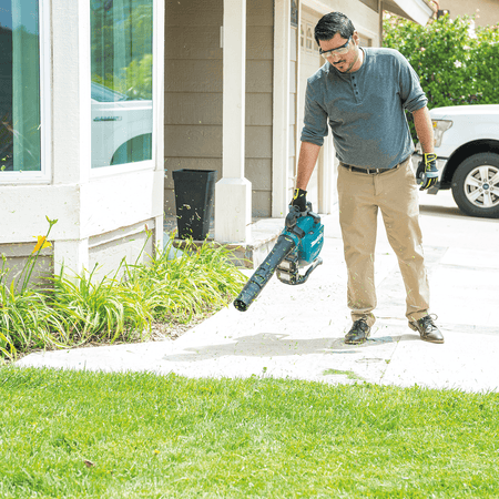 Worker clearing grass clippings and debris off a residential concrete driveway with a Makita blower.