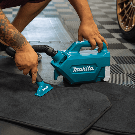 Operator using the Makita XLC07Z cordless canister vacuum with a wide nozzle to clean dirt and debris off a rubber car floor mat.