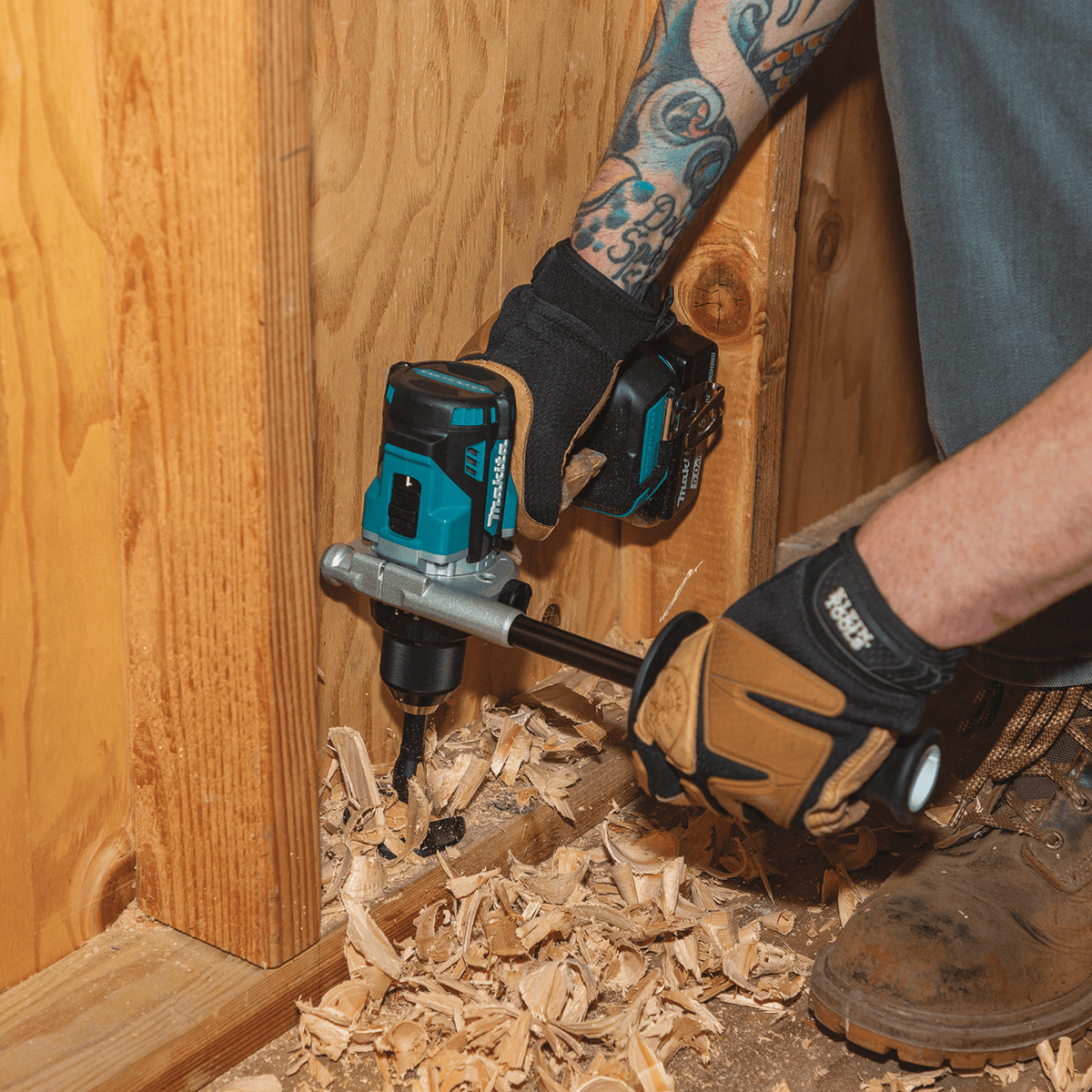Worker drilling downward through a bottom wall plate with the Makita XPH14 hammer drill using a spade bit and side handle for stability.