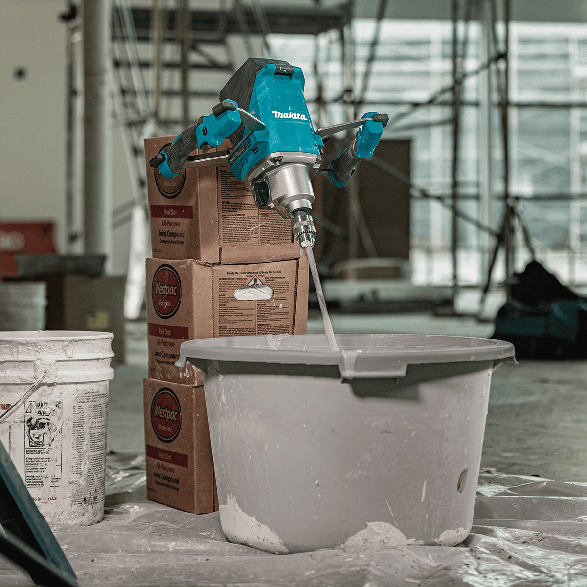 Makita 40V XGT cordless mixer resting upright near stacked boxes of joint compound on an active commercial jobsite.