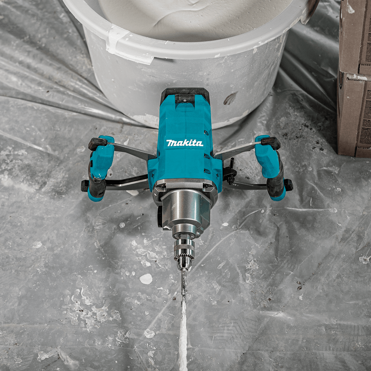 Top-down view of the Makita 40V XGT cordless mixer resting on a drop cloth next to a bucket of freshly mixed compound.