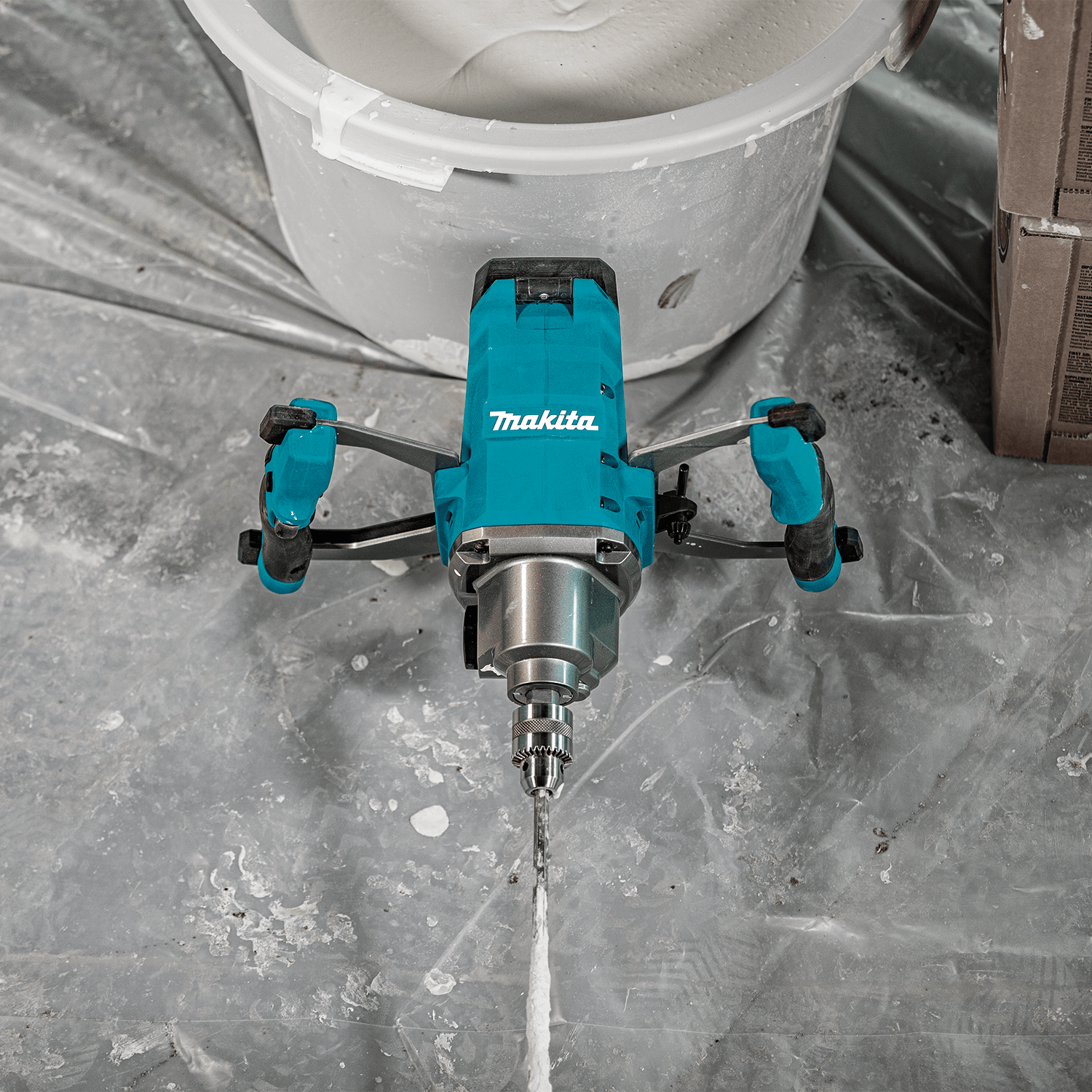 Top-down view of the Makita 40V XGT cordless mixer resting on a drop cloth next to a bucket of freshly mixed compound.
