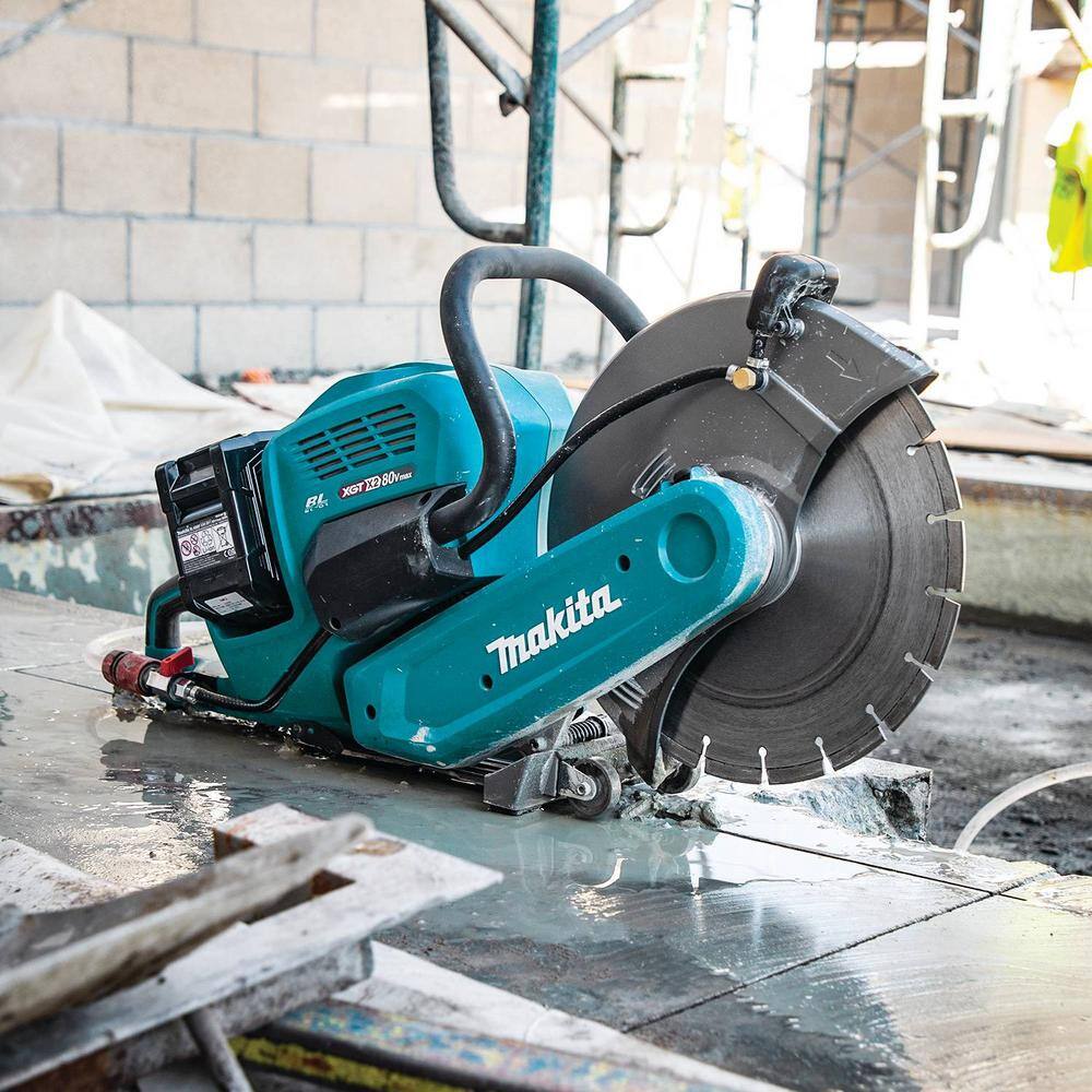 Low-angle profile view of the Makita 14 inch cordless power cutter saw sitting idle on a wet concrete floor pad inside a commercial building under construction, showcasing its built-in dual floor-tracking guide wheels.