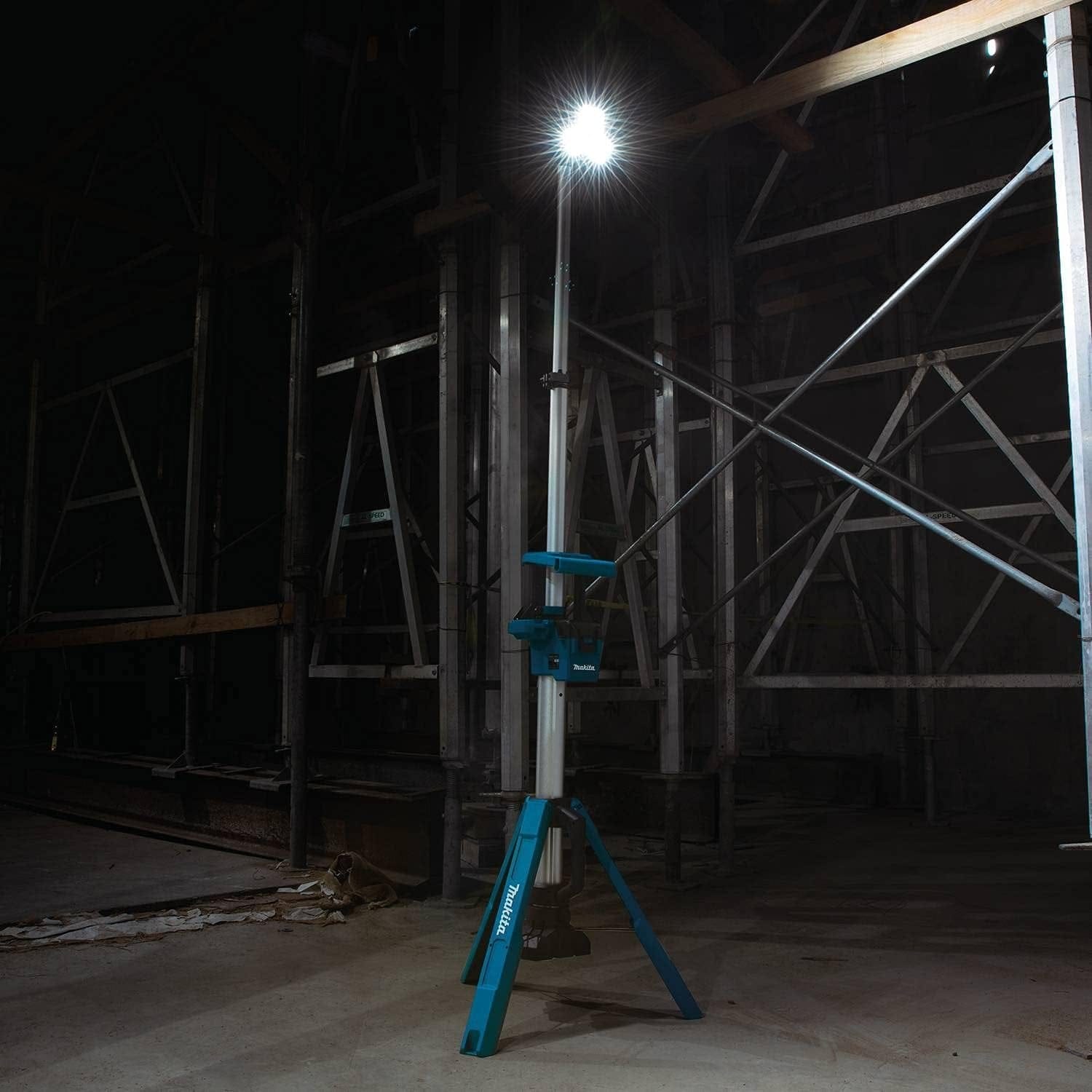 Action shot of the Makita DML813 18V LXT tower light fully extended and powered on in a dark industrial scaffolding area, providing high-intensity overhead LED illumination.