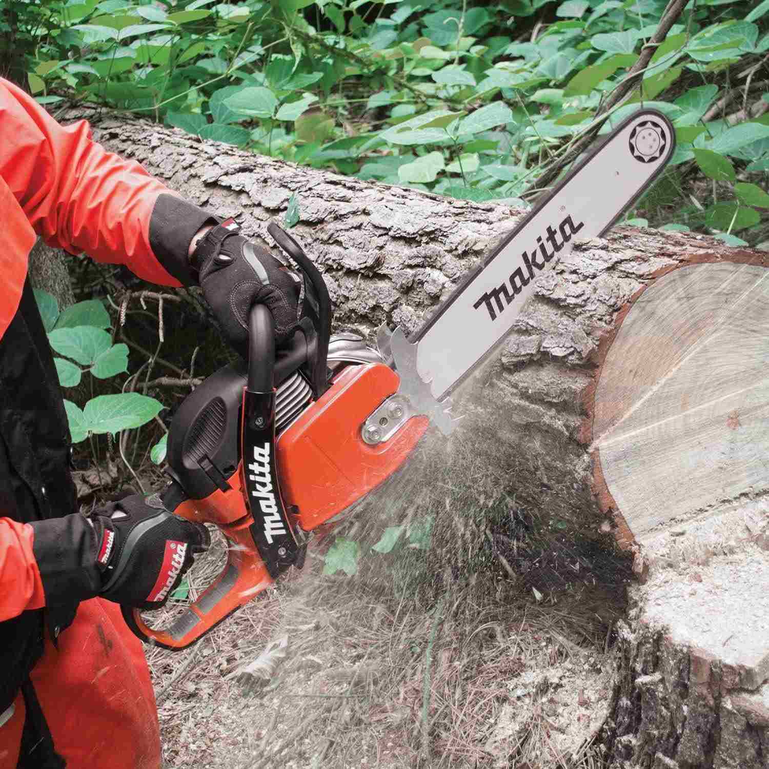 A user wearing orange protective safety gear cuts through a large fallen log with the Makita chainsaw, creating a spray of sawdust.