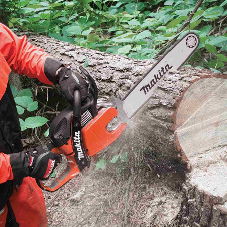 A user wearing orange protective safety gear cuts through a large fallen log with the Makita chainsaw, creating a spray of sawdust.