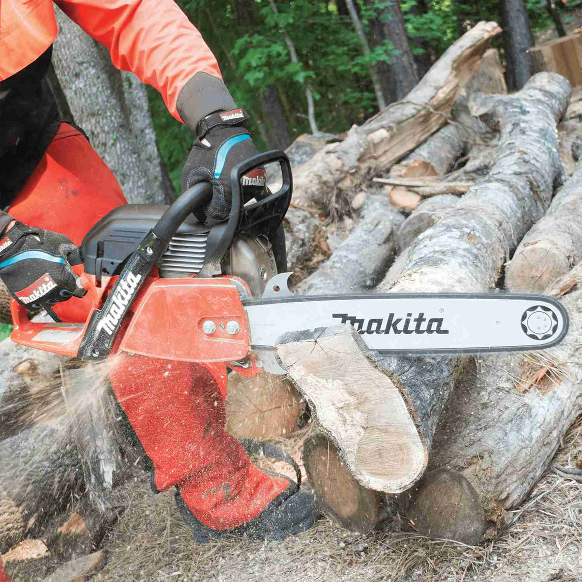 A user wearing orange protective safety gear cuts through a large fallen log with the Makita chainsaw, creating a spray of sawdust.