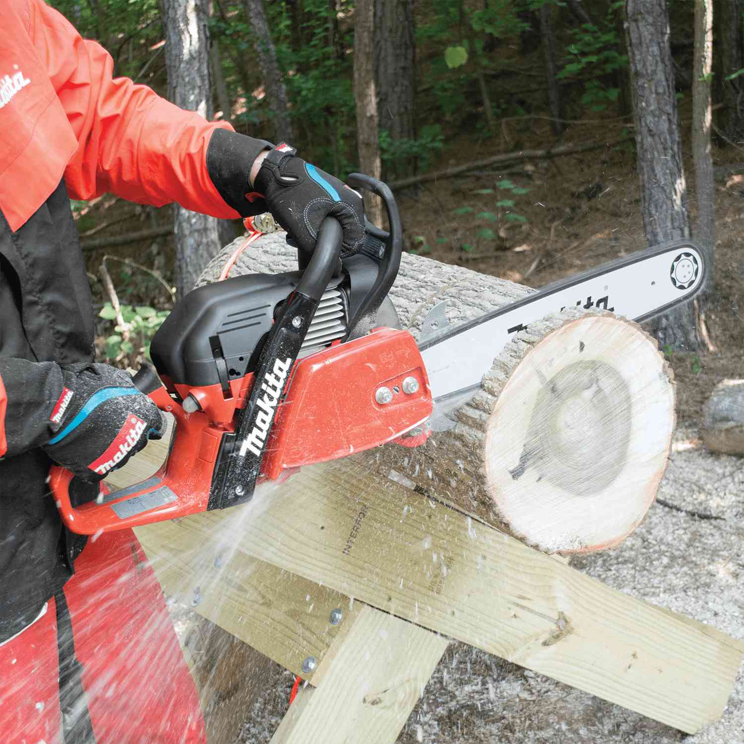 A user wearing orange protective safety gear cuts through a large fallen log with the Makita chainsaw, creating a spray of sawdust.