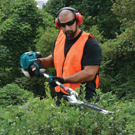 Action shot of a landscaper wearing an orange safety vest and ear protection using the Makita articulating hedge trimmer to cut the flat top of a hedge.