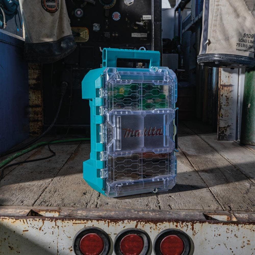 A Makita MAKTRAK T-90065 organizer stood vertically on a jobsite surface, showcasing its robust comfort-grip carrying handle and the visibility provided by the clear lid.