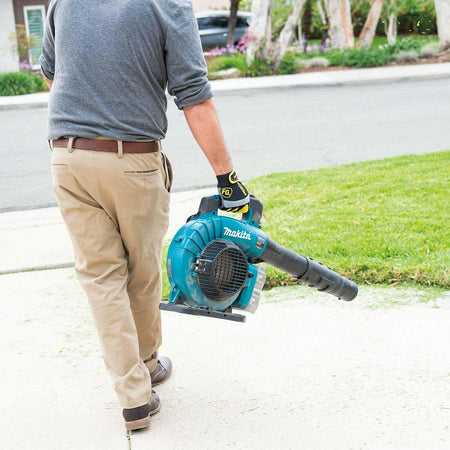 Rear view of a user carrying the Makita XBU04Z blower along a concrete path, holding it by the top handle. The inlet grate and attached nozzle are visible.