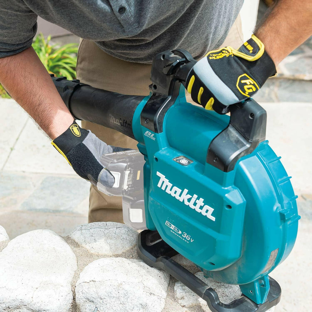 A user wearing work gloves loads an 18V LXT battery into the Makita XBU04Z brushless blower, resting the unit on a low stone wall. The telescoping nozzle is attached.