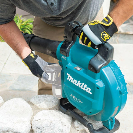 A user wearing work gloves loads an 18V LXT battery into the Makita XBU04Z brushless blower, resting the unit on a low stone wall. The telescoping nozzle is attached.