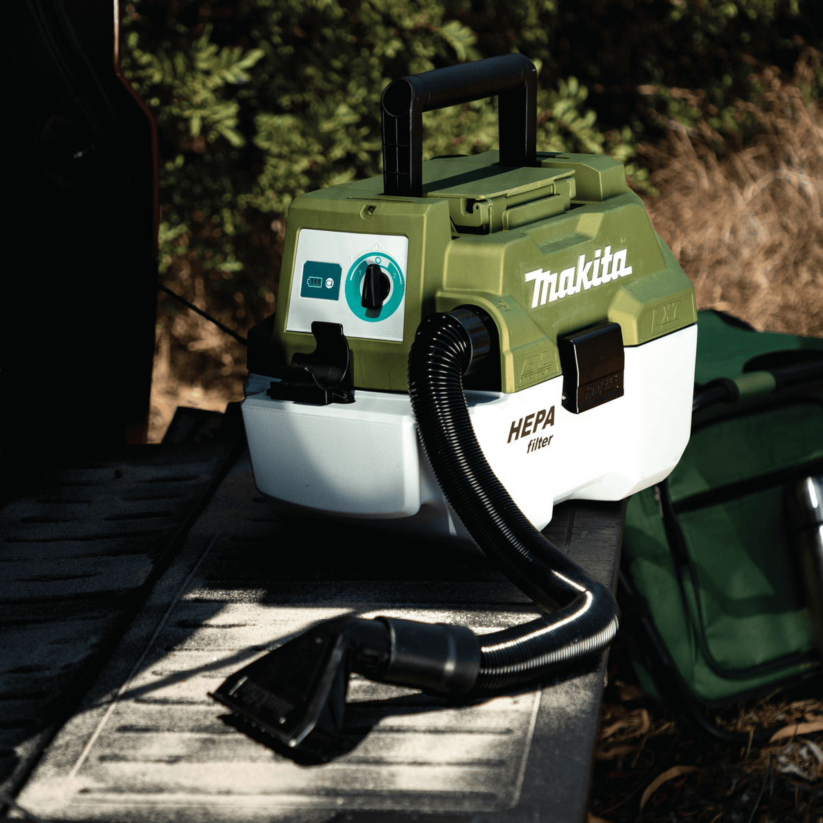 Makita ADCV11Z 18V LXT wet dry vacuum sitting on a vehicle tailgate equipped with a flexible anti-static hose and wide upholstery nozzle.