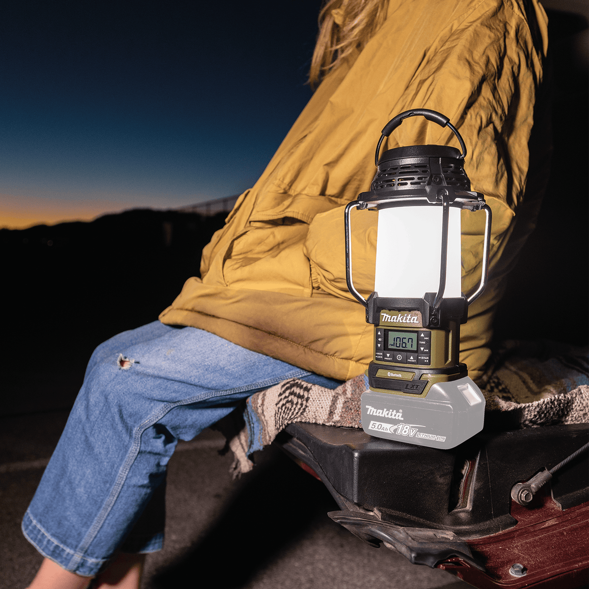 Camper sitting on a vehicle tailgate at night illuminated by the 360-degree ambient glow of the Makita ADRM13 18V LXT lantern.