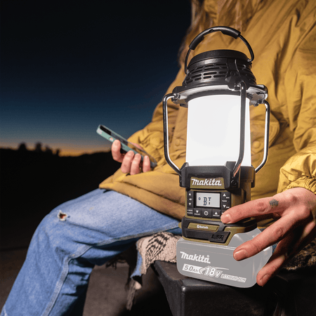 Person adjusting the Bluetooth volume and media control buttons on the Makita ADRM13 18V LXT lantern during an evening campsite gathering.