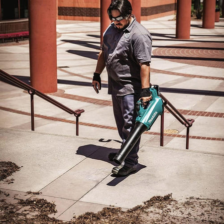 Full view of a landscaping professional wearing a uniform and protective safety glasses operating the Makita 18V LXT cordless handheld blower in an outdoor commercial plaza. The worker carries the lightweight lawn cleaner down a set of concrete stairs to sweep away matted yard debris.