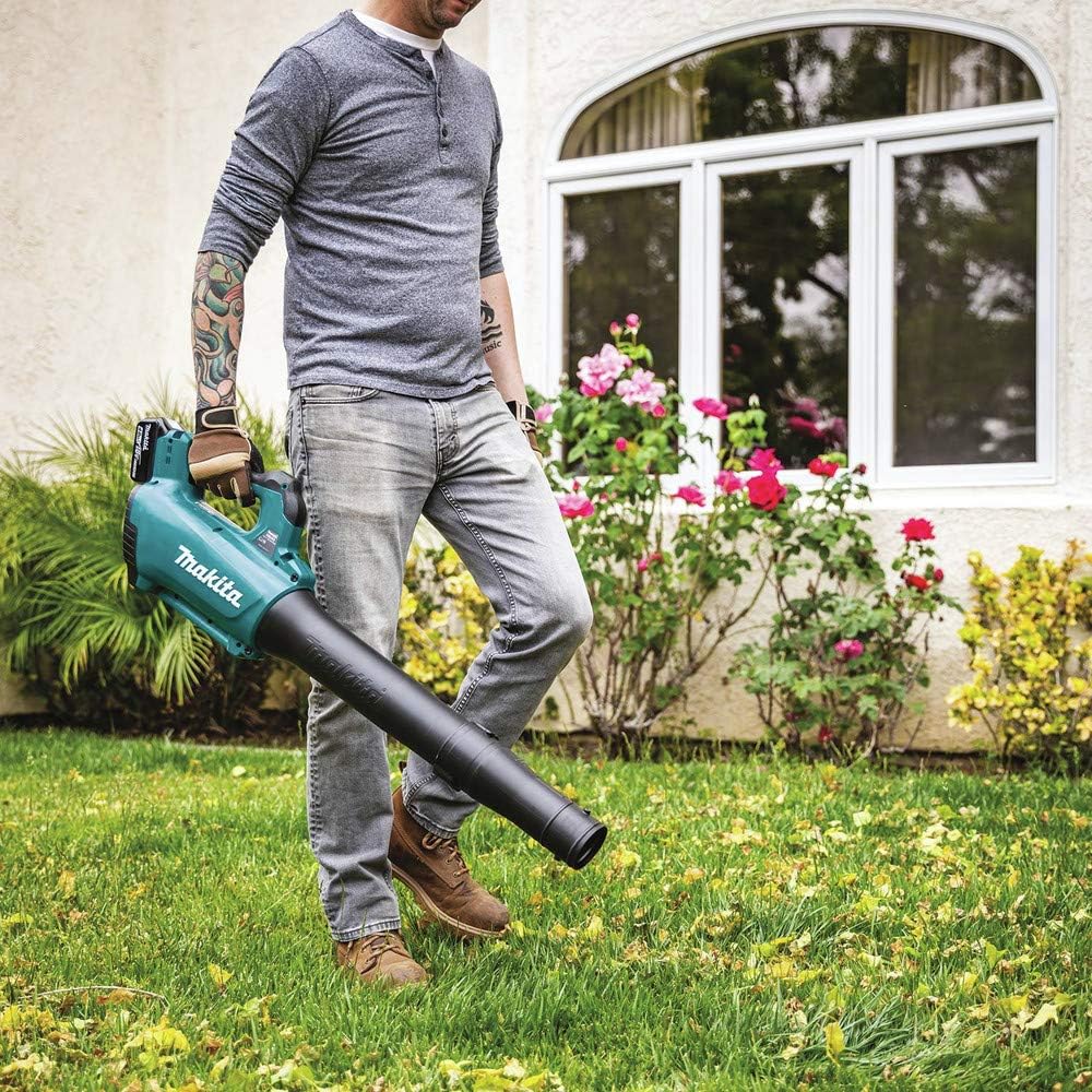 A man in a long-sleeve shirt and jeans walks across a green residential lawn using the teal Makita 18V LXT cordless handheld blower. The garden power tool is pointed downward to clear scattered leaves and lawn debris near flowerbeds and a house window.