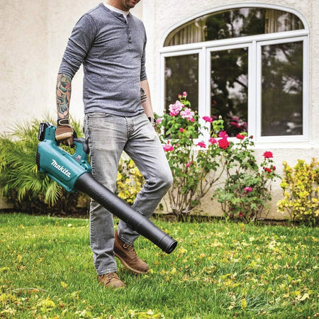 A man in a long-sleeve shirt and jeans walks across a green residential lawn using the teal Makita 18V LXT cordless handheld blower. The garden power tool is pointed downward to clear scattered leaves and lawn debris near flowerbeds and a house window.