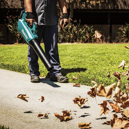 An outdoor action shot of a person using the handheld Makita brushless leaf blower to clear dry, brown leaves off a concrete walkway. The yard debris is shown swirling into the air from the powerful 459 CFM velocity generated by the cordless lawn cleaner.
