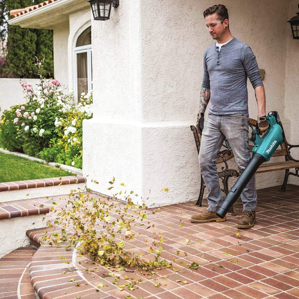 A man using the lightweight Makita cordless garden blower to clean a brick patio porch area next to a house. The variable speed blower tube is directed toward a pile of autumn leaves, sending them flying off the tiled outdoor surface.