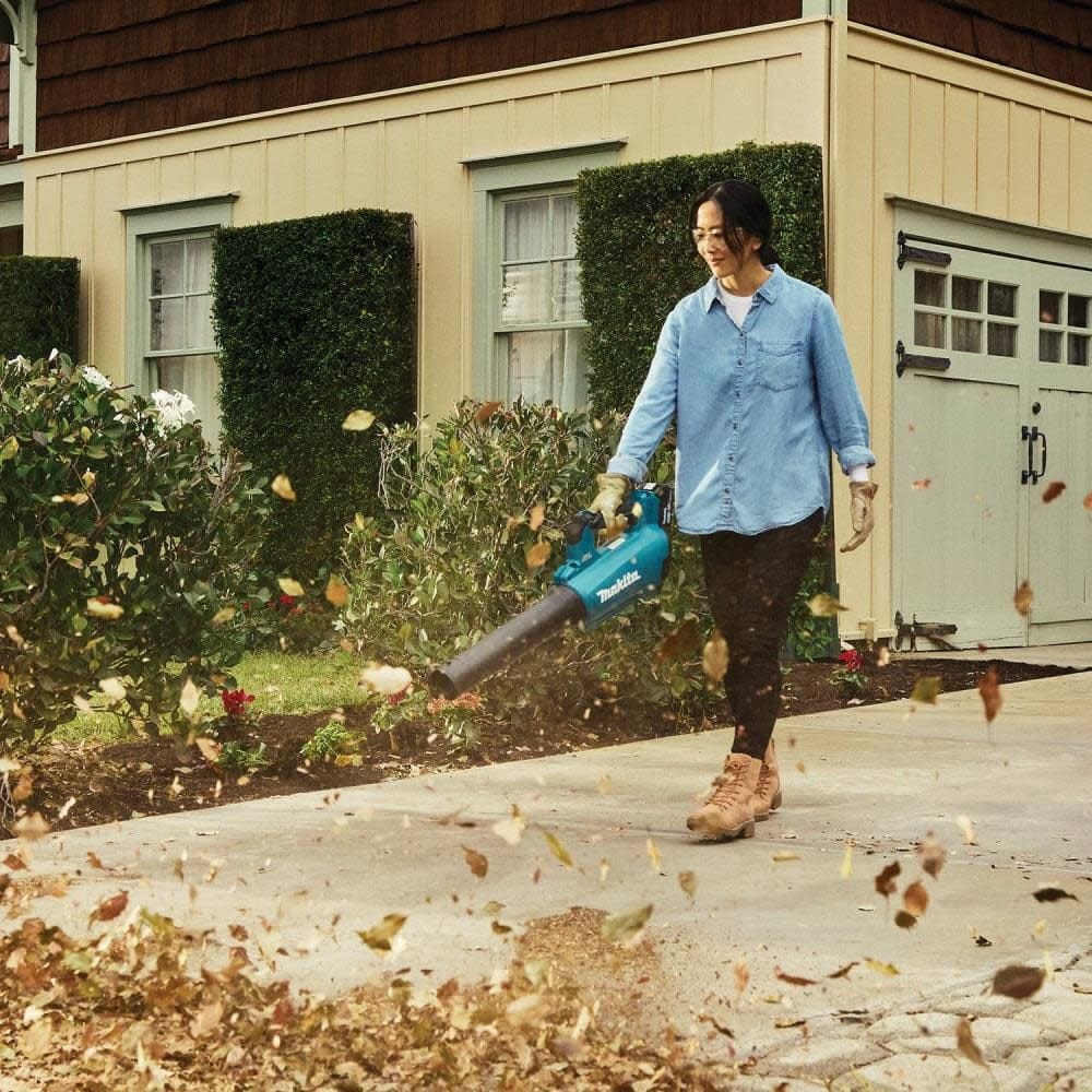 A woman wearing a denim shirt and work gloves operates the Makita 18V LXT handheld leaf blower to sweep a concrete driveway. A large cloud of dry fall leaves is blown forward by the high-velocity stream from the teal cordless power tool.