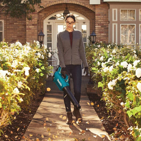 A woman wearing safety glasses and a jacket uses the teal Makita brushless leaf blower to clear a stone tile sidewalk. She is surrounded by white rose bushes as the powerful handheld yard cleaner sweeps away falling autumn leaves.