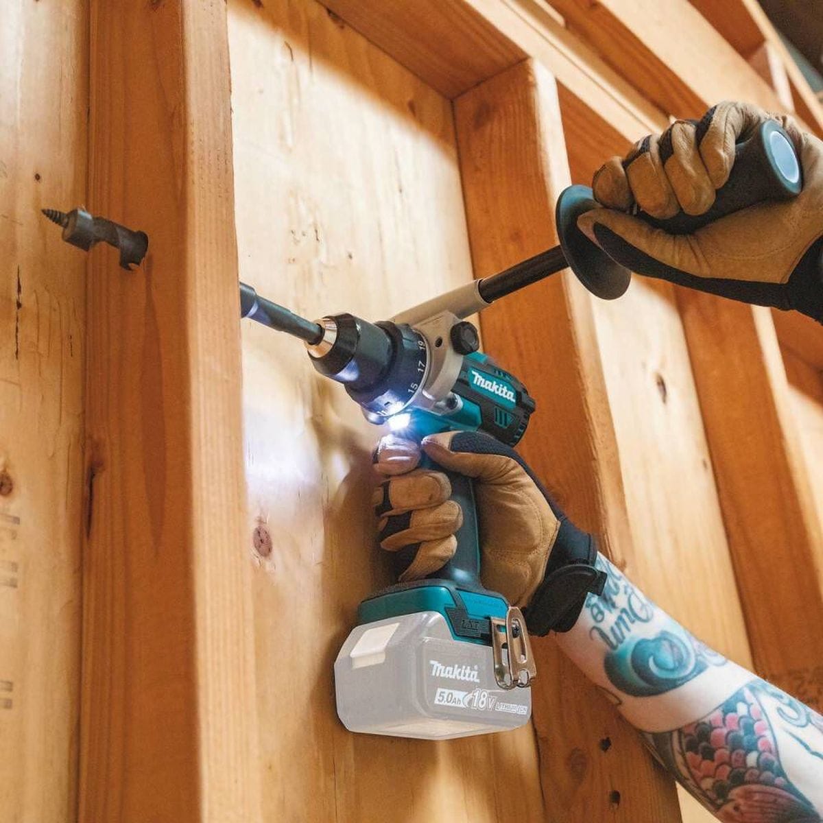 A jobsite application shot showing the Makita 18V LXT brushless hammer drill boring through a vertical wall stud, highlighting the integrated L.E.D. work light and the ergonomic use of the auxiliary side handle for stability.