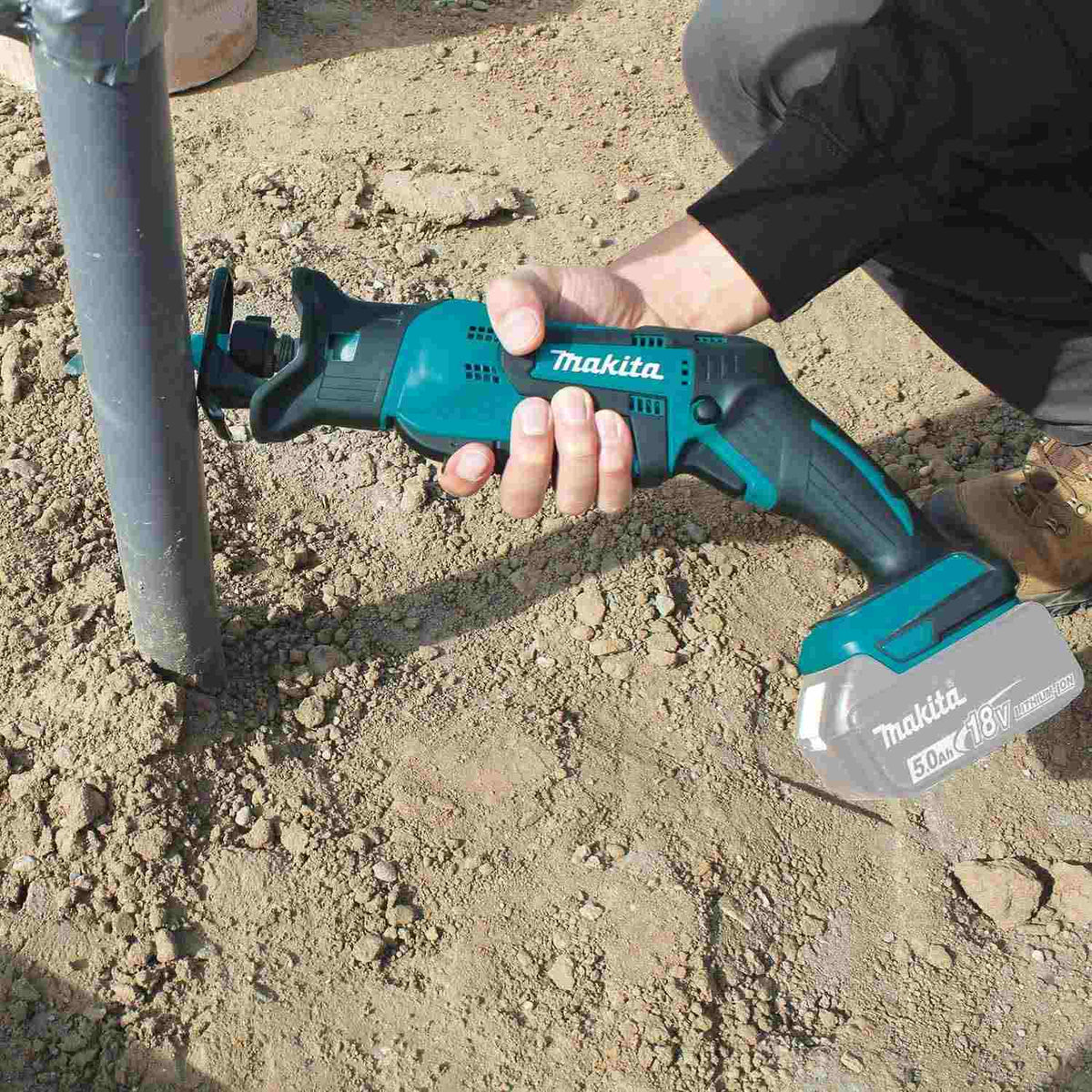 A worker using the Makita XRJ01Z 18V LXT cordless reciprocating saw with a battery attached to efficiently cut through a vertical PVC pipe close to the dirt ground.