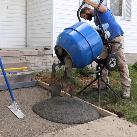An action photograph of a professional worker using the Marshalltown MIX3 electric mixer to pour freshly mixed concrete directly into a sidewalk form, demonstrating the convenience of the support pedestal.