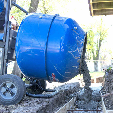 A close-up action shot of the Marshalltown MIX3 3 cubic foot mixer drum tilted forward, showing wet concrete pouring into a trench, highlighting the mixer's efficiency for residential and commercial projects.
