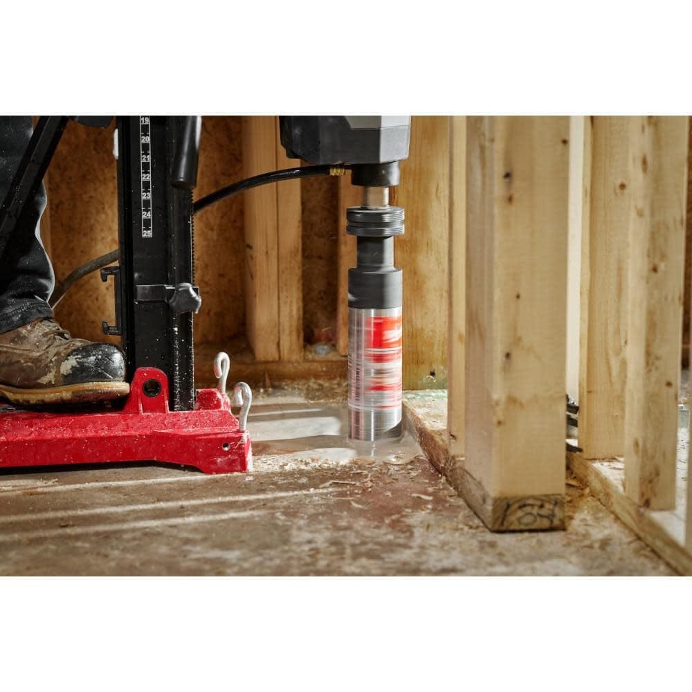 A close-up action shot of a rig-mounted Milwaukee wet core bit boring a hole through a concrete floor inside a residential wood-framed building.