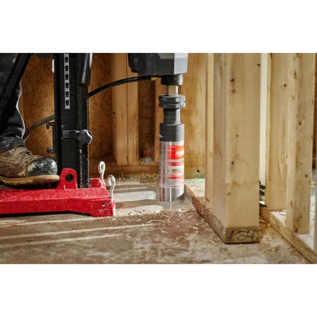 A close-up action shot of a rig-mounted Milwaukee wet core bit boring a hole through a concrete floor inside a residential wood-framed building.