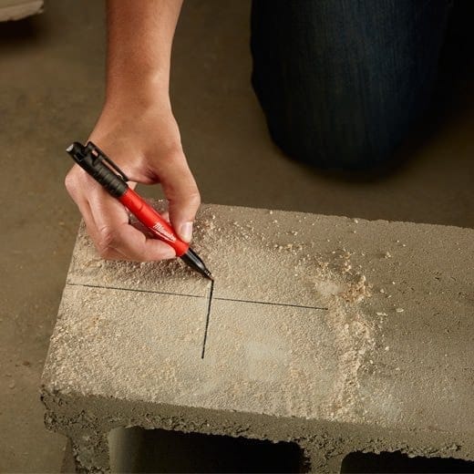 High-angle action shot of a hand using a red Milwaukee INKZALL marker to draw black layout lines on a rough concrete cinder block.