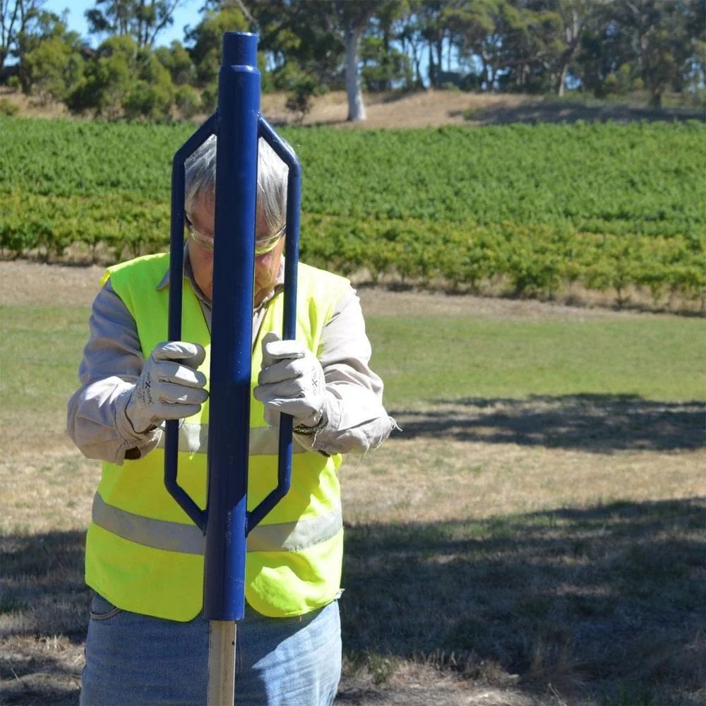 Operator wearing high visibility safety gear holding the blue star dropper hammer vertically to drive a post in a grassy field.