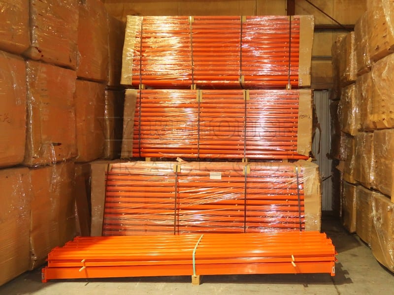 A warehouse interior showing large, bundled stacks of new 120-inch industrial safety orange teardrop pallet rack load beams wrapped in protective plastic for distribution.