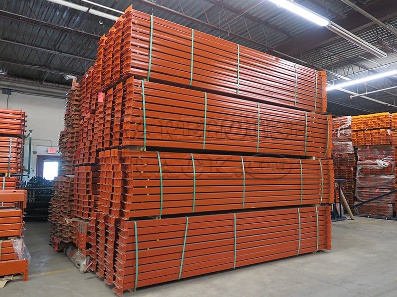 An angled perspective shot of multiple bundles of new 12-foot safety orange teardrop pallet rack beams in a warehouse, highlighting the roll-formed steel construction and shaved-pin end connectors.