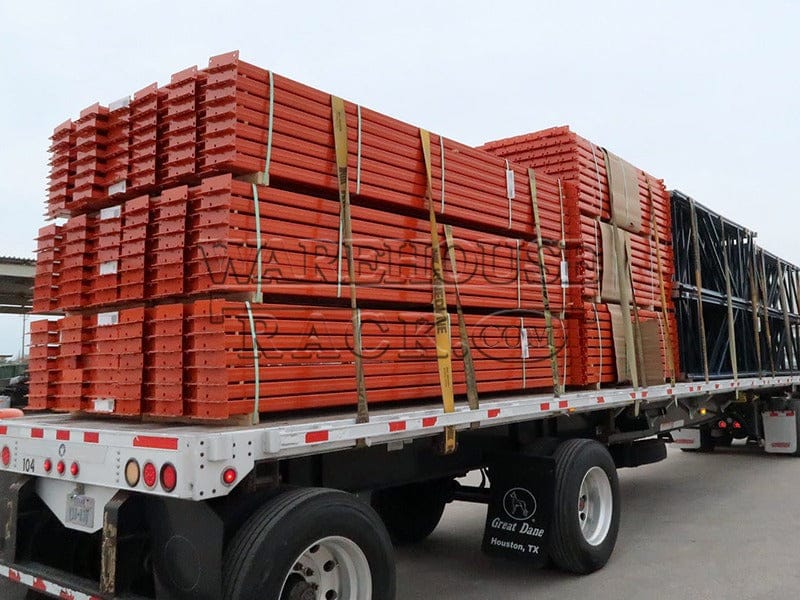 A side-profile view of multiple bundles of new 144-inch industrial safety orange teardrop pallet rack load beams secured on a flatbed trailer for delivery to a warehouse or distribution center.
