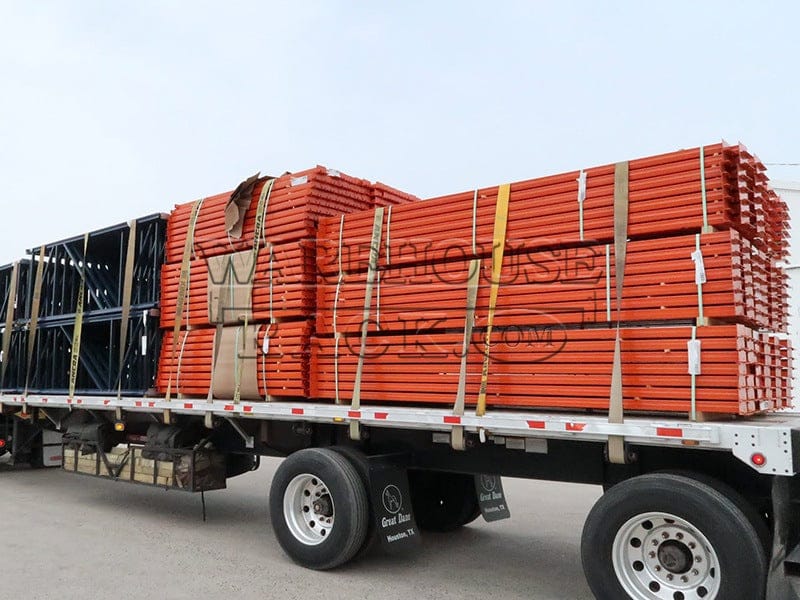 A wide perspective view of a transport truck loaded with bundles of safety orange 12-foot warehouse load beams and blue selective pallet rack upright frames, ready for industrial storage installation.