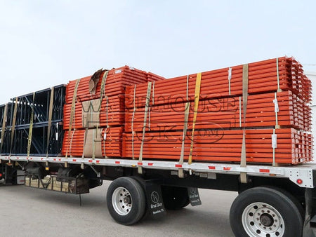 A wide perspective view of a transport truck loaded with bundles of safety orange 12-foot warehouse load beams and blue selective pallet rack upright frames, ready for industrial storage installation.