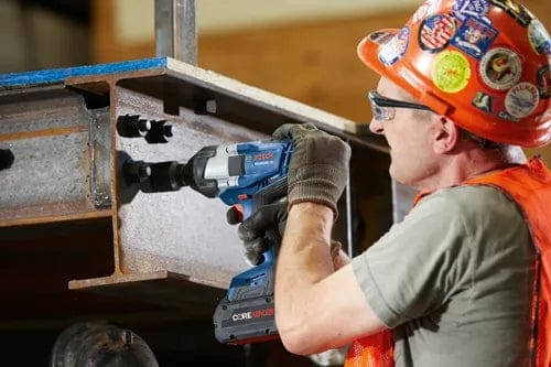 Construction worker drilling into a steel I-beam with a Bosch cordless drill, wearing safety gear including hard hat, vest, and gloves.