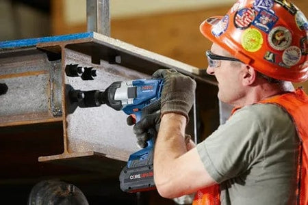 Construction worker drilling into a steel I-beam with a Bosch cordless drill, wearing safety gear including hard hat, vest, and gloves.