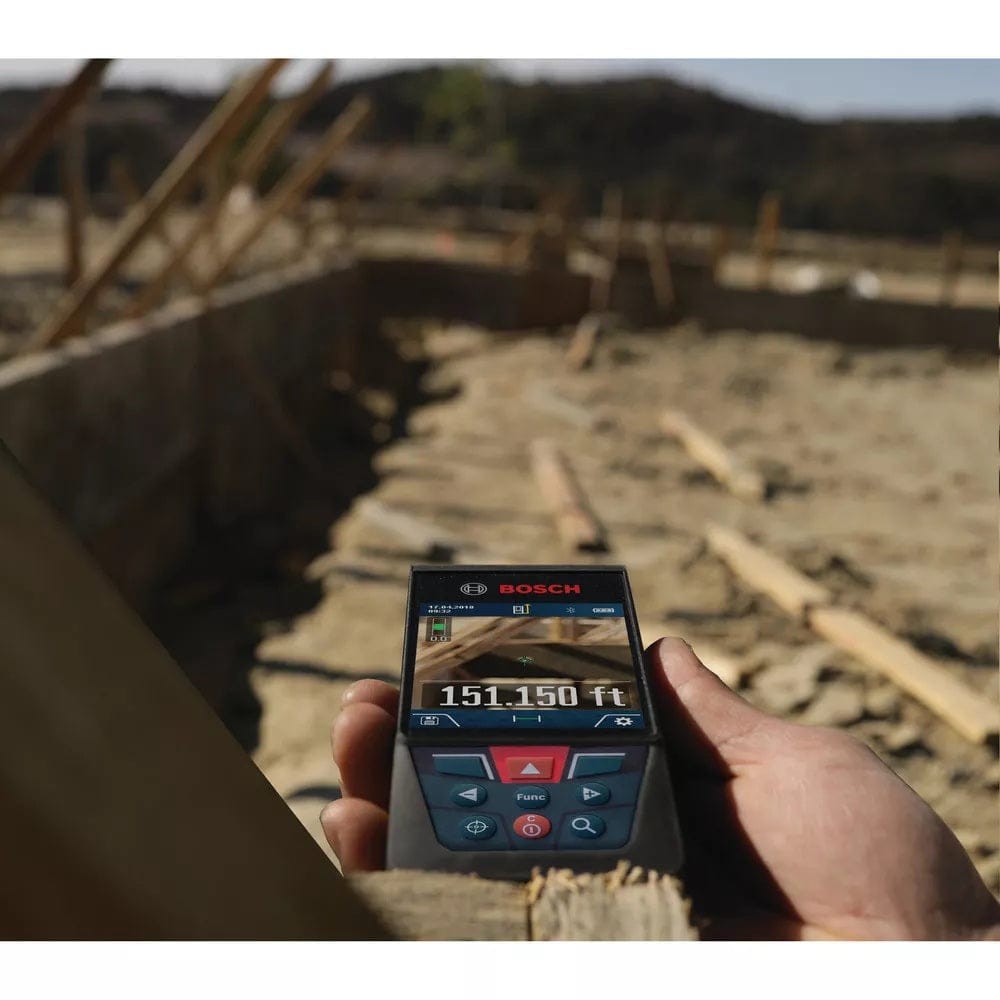 Hand holding a Bosch laser distance measurer at a construction site. The screen shows a reading of 151.150 feet. Background includes wooden framing and dirt, indicating early-stage building work.