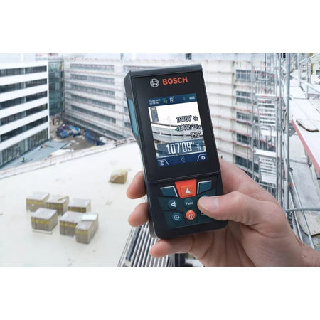Close-up of Bosch GLM 50 C laser distance measurer held at a construction site. The screen displays multiple measurements including angles and distances, with scaffolding and buildings in the background.