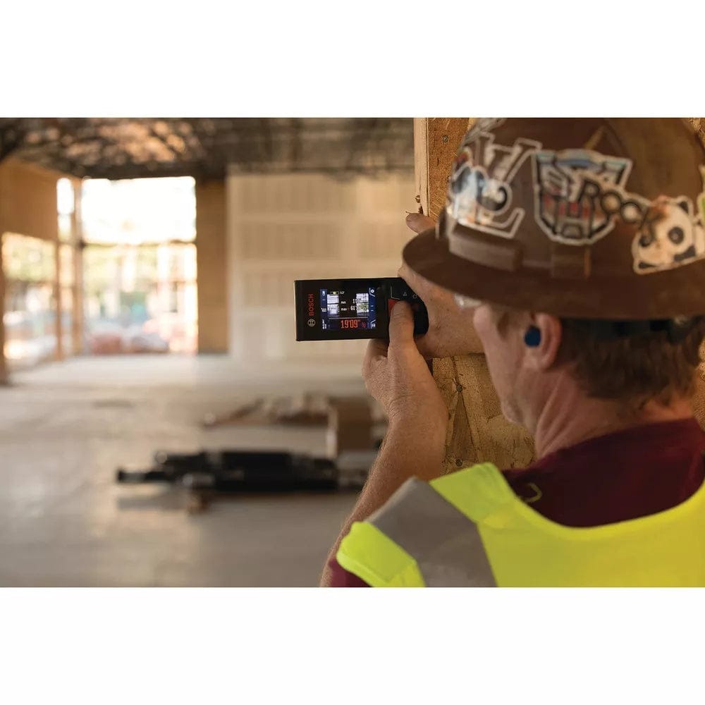Construction worker wearing a hard hat and safety vest uses a Bosch laser distance measurer inside a partially built structure. The device is aimed at a wall, and its screen displays measurement data. The background includes exposed framing and construction materials.