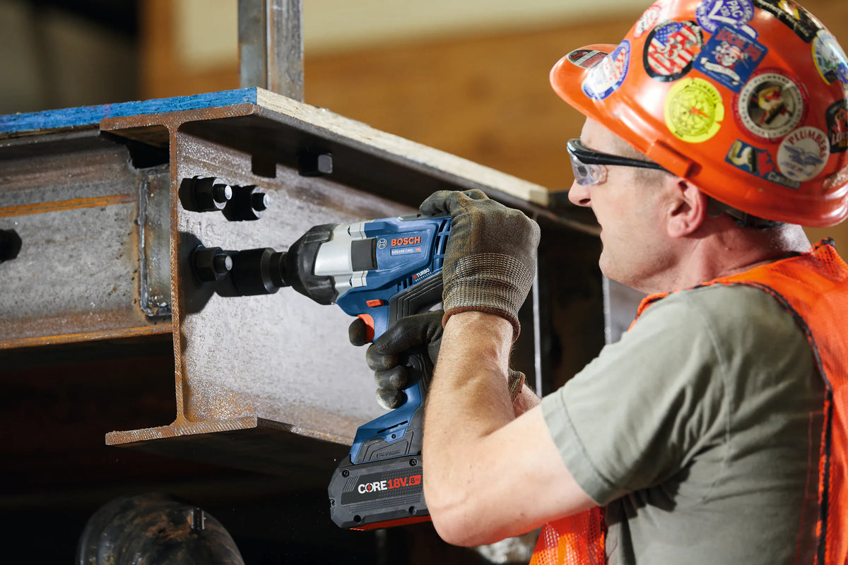An on-site application shot of a structural ironworker wearing a stickered orange hard hat and safety glasses using the Bosch 18V PROFACTOR impact wrench to fasten heavy-duty structural steel bolts onto an industrial I-beam.