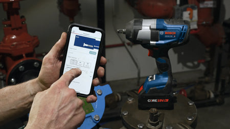 A close-up demonstration shot showing an operator using a smartphone to configure custom parameters on the Bosch Toolbox mobile app interface alongside the connected-ready 18V impact wrench.